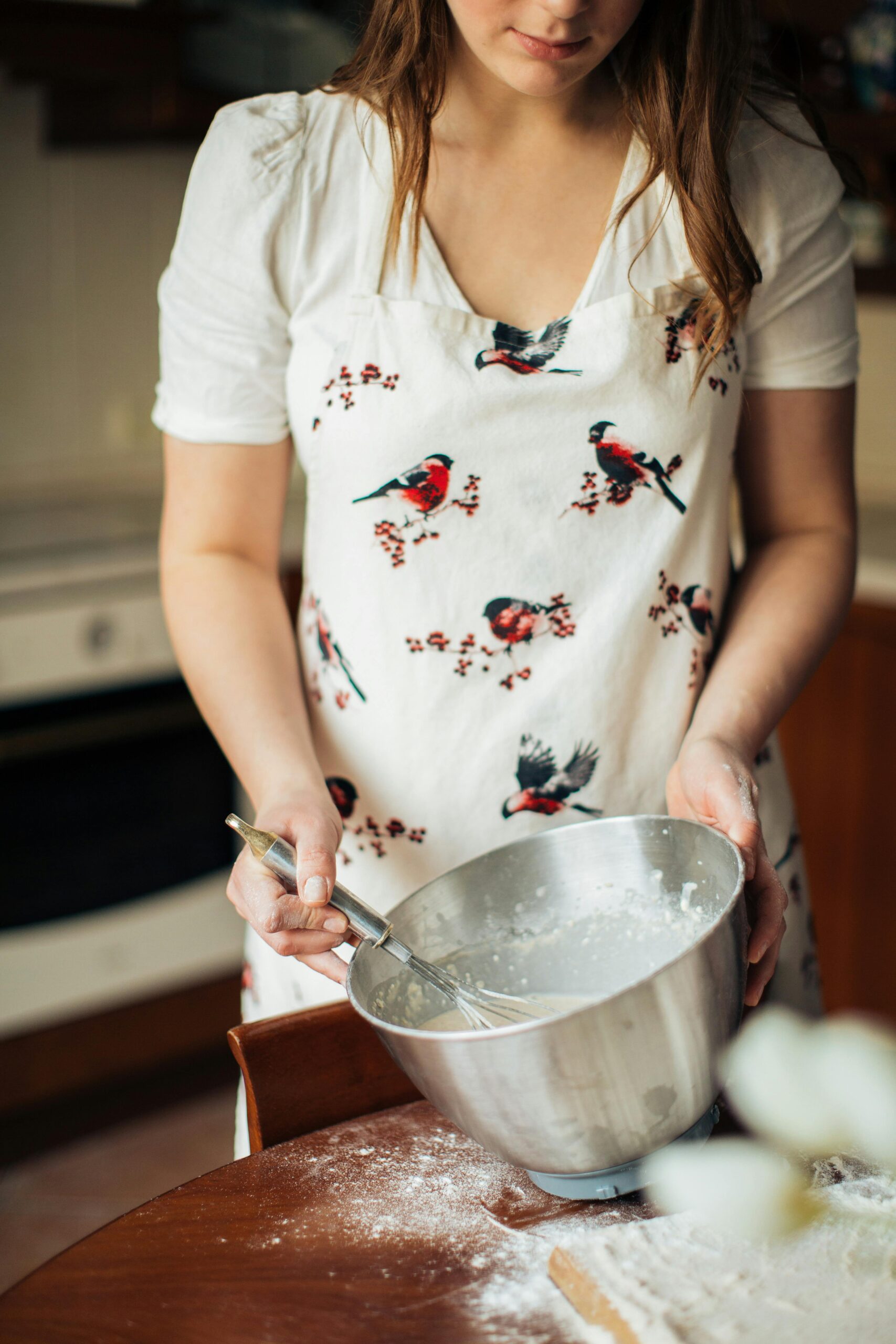 A woman in a kitchen mixing dough in a bowl while wearing a bird-patterned apron.